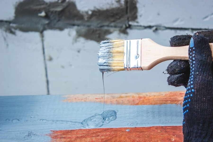 Main gantée tenant un pinceau chargé de matière grise pour montrer comment appliquer une peinture à base d'algues sans laisser de traces sur du bois.