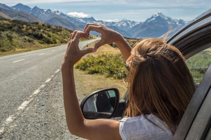 Une voyageuse faisant un signe de cœur avec ses mains par la fenêtre d'une voiture, devant un paysage de montagnes enneigées lors de la planification d'un road trip.