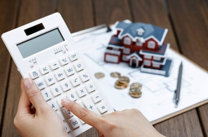 Calculatrice blanche tenue en main devant une maquette de maison et des pièces de monnaie pour évaluer le pouvoir d achat primo accédant france.