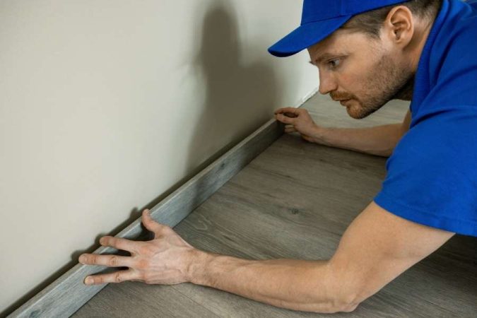 Un artisan installe avec précision une plinthe en bois grise le long d'un mur dans une pièce rénovée.